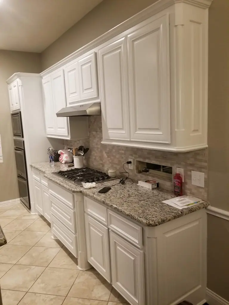 A modern white wet bar featuring glass cabinets, black pulls, a wine cooler, and a diamond-pattern tile backsplash by Texas Star Painting of Austin.