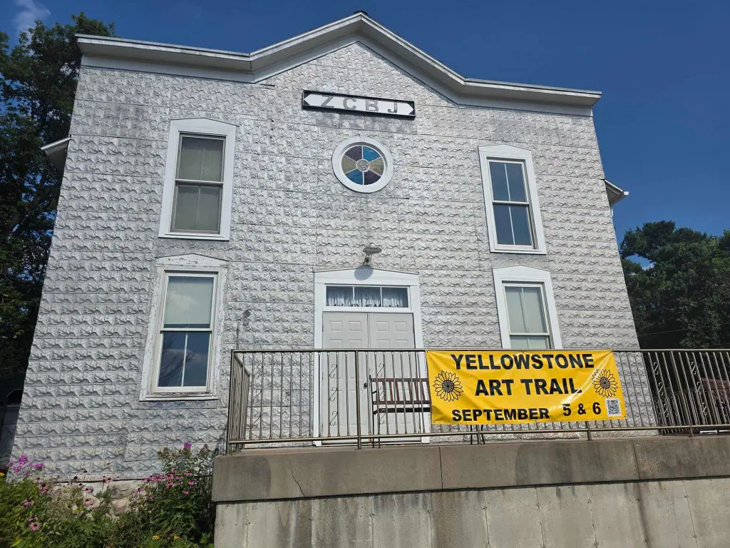 The historic, textured white facade of a two-story hall, featuring a round stained glass window and a yellow 'Yellowstone Art Trail' banner, awaits its fresh coat of professional care from Texas Star Painting of Austin.