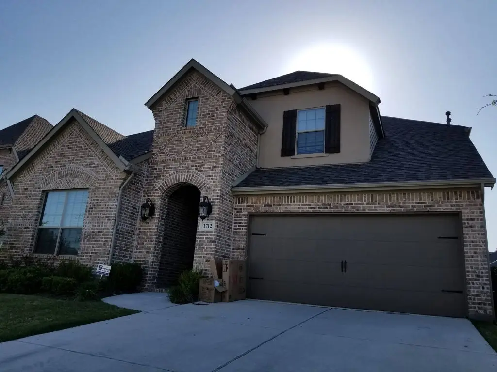 Two-story brick house with a dark shingled roof, a brown garage door, dark window shutters, and a covered arched entrance, with some cardboard boxes stacked near the front door.