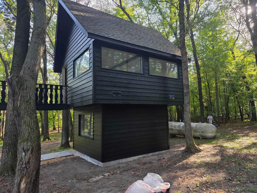 Black modern forest retreat with high windows, a deck, and an assortment of autumn pumpkins.