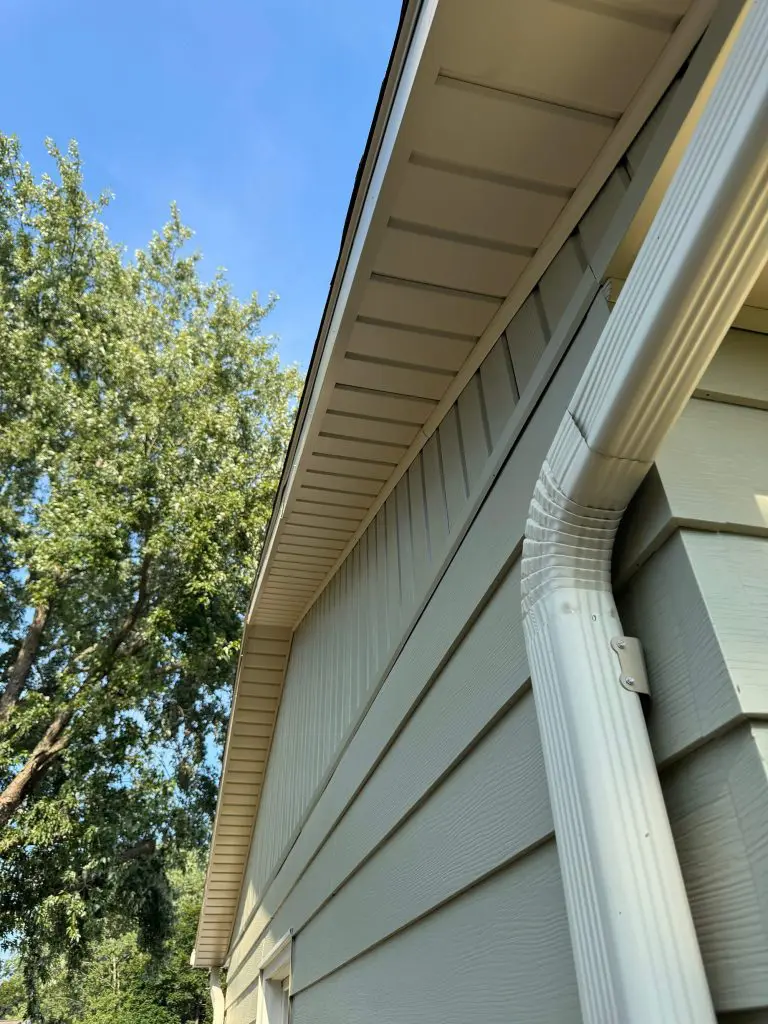 Light olive or pale sage green horizontal lap siding on a Texas home with matching vertical siding on the gable end, featuring white fascia, soffit, and a white downspout against a blue sky.