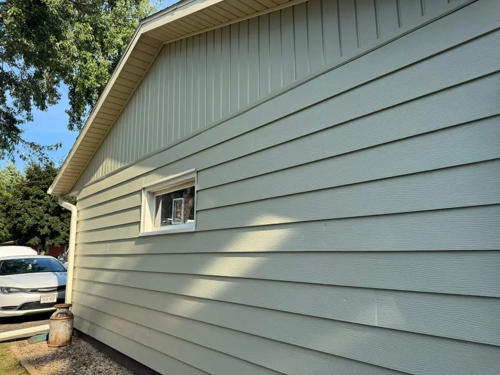 Light sage green or pale olive-colored horizontal lap siding on a Texas house with a small, white-trimmed window, tan vertical siding on the gable end, and a white sedan parked nearby.