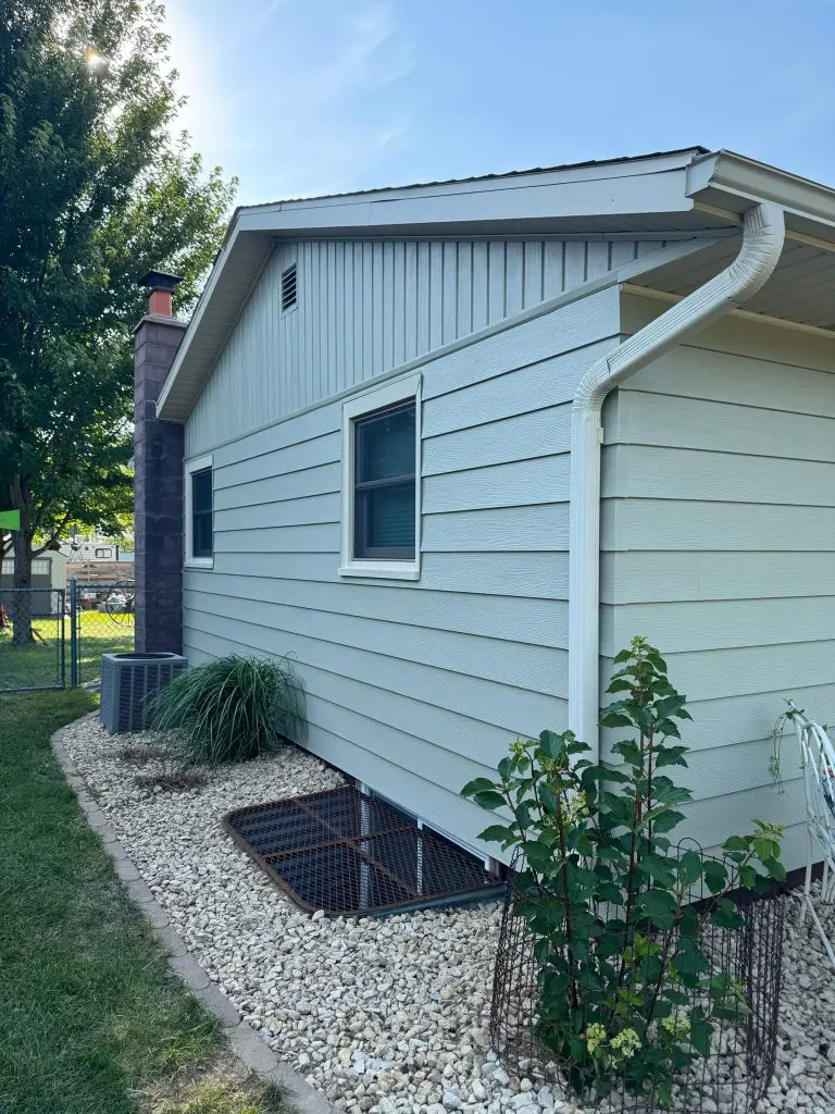 Light blue-gray siding on a Texas home's exterior, featuring two white-trimmed windows, a dark stone chimney, a gutter downspout, and a rock landscaping bed with foundation plantings.