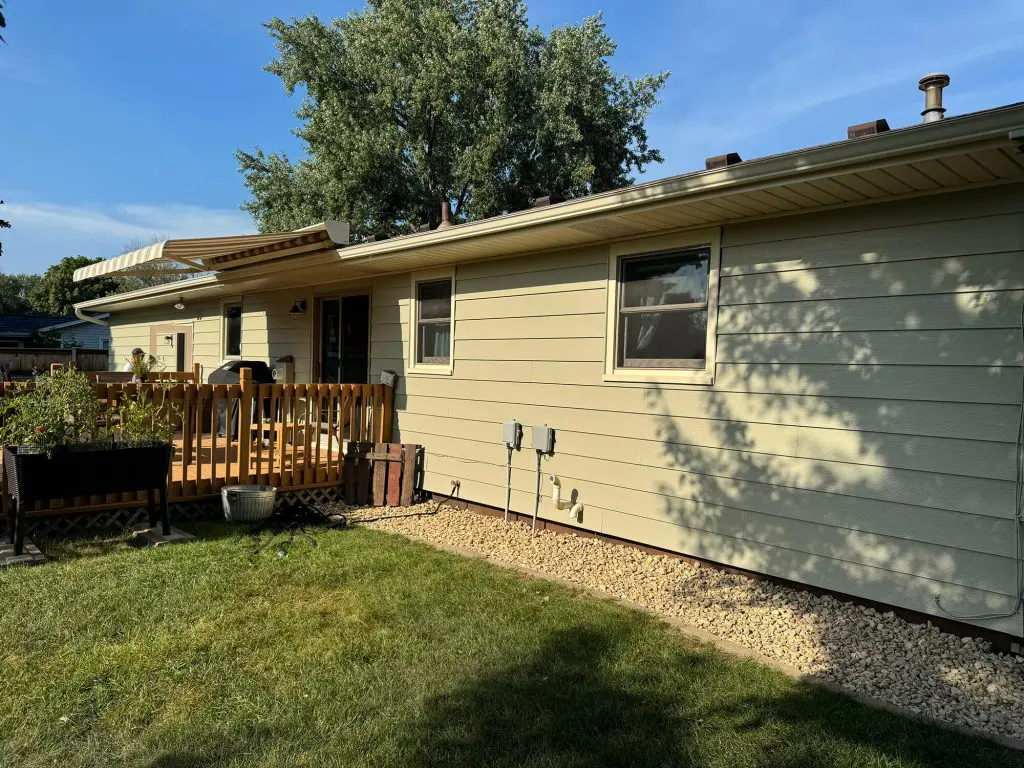 One-story home with light beige siding, a raised wooden deck with a low railing and a striped retractable awning, and a gravel border along the foundation next to a green lawn under a sunny blue sky.