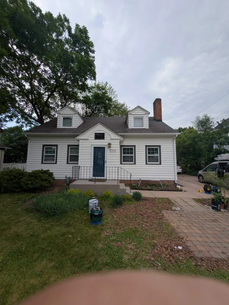 White clapboard cottage with a dark blue front door and porch railing, two small dormers, a red brick chimney, and surrounding landscaping, photographed on an overcast day.