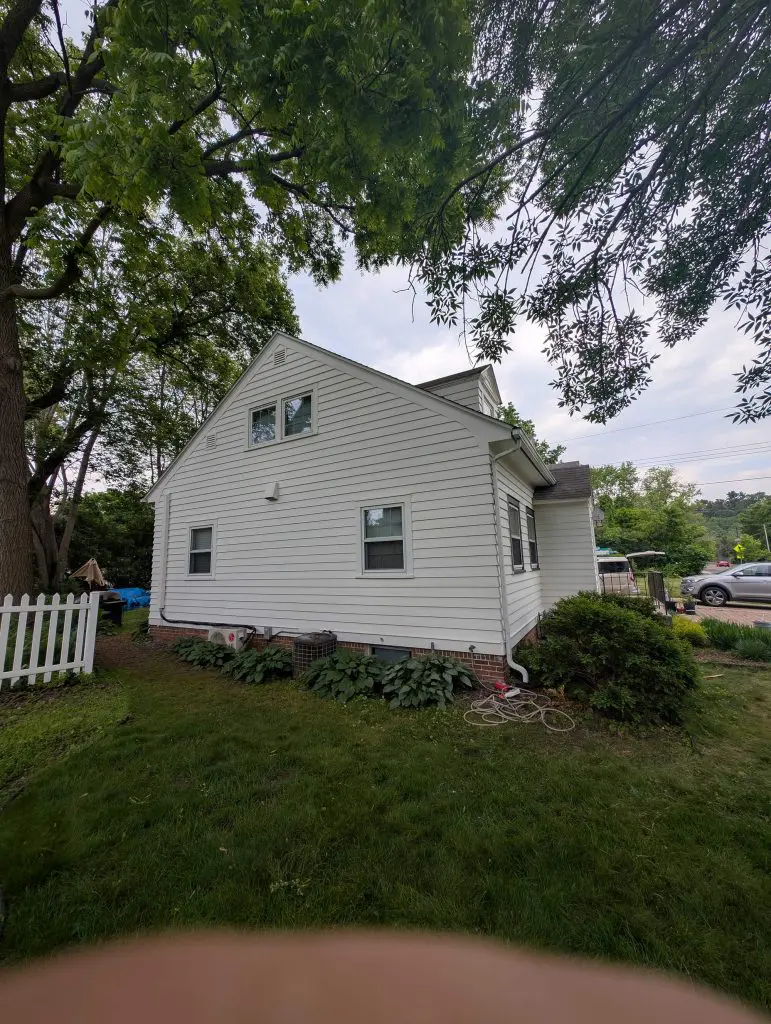 White clapboard-sided house with a steep gable roof, a small white picket fence, large trees overhead, shrubs and hostas planted along the foundation, and two air conditioning units.