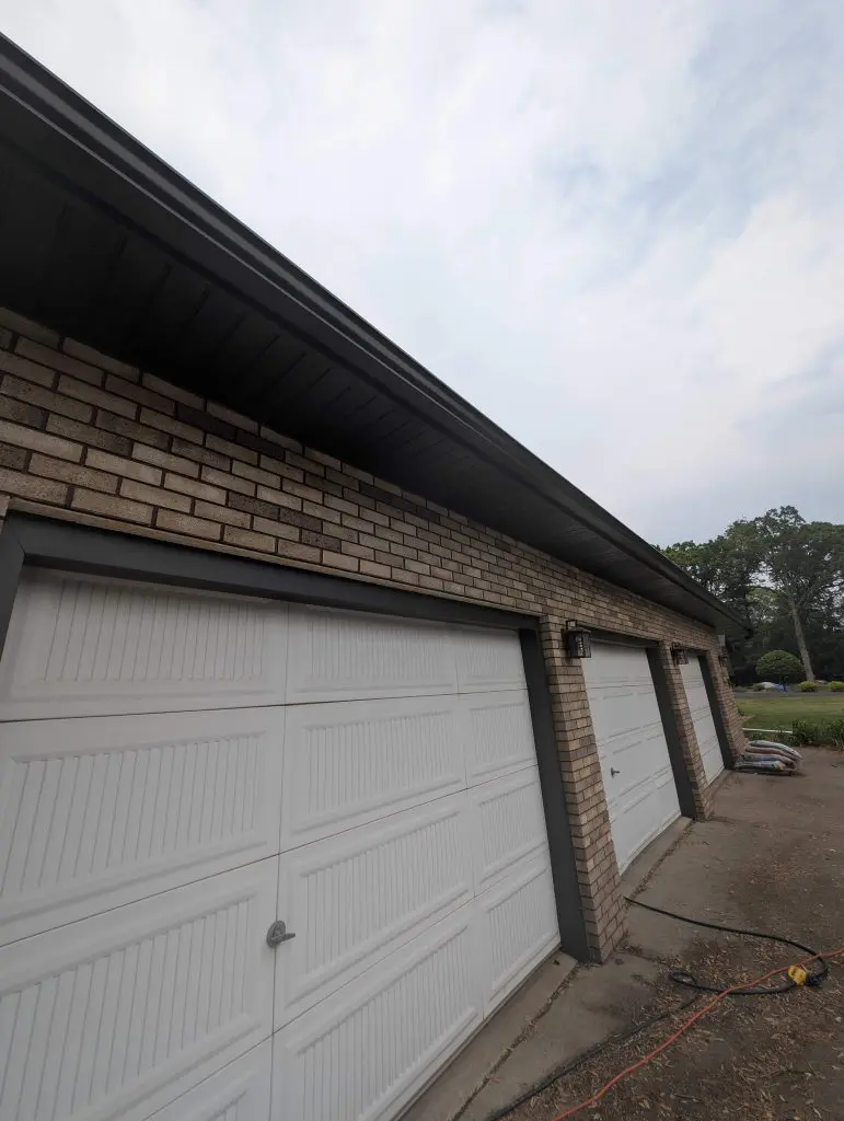 Three side-by-side white panel garage doors set in a tan brick wall with dark brown trim and gutters, photographed from a low-angle perspective under an overcast sky.