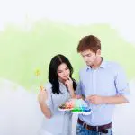 A couple reviewing paint color swatches in front of a partially painted wall, considering the signs it's time to repaint interior walls in their home.