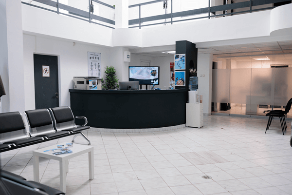 Modern, clean, and bright medical or corporate lobby with a curved black reception desk, a central monitor displaying information, white tiled floors, metal-framed waiting chairs, and a water cooler.
