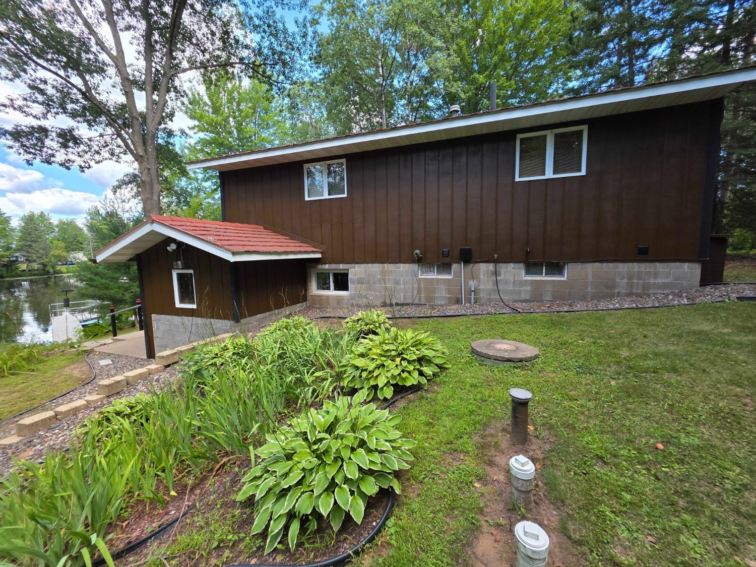 The back of a brown, two-story home with a red-roofed addition, nestled near a wooded lakefront and accented by vibrant green landscaping, expertly painted by Local Painters in Cedar Park