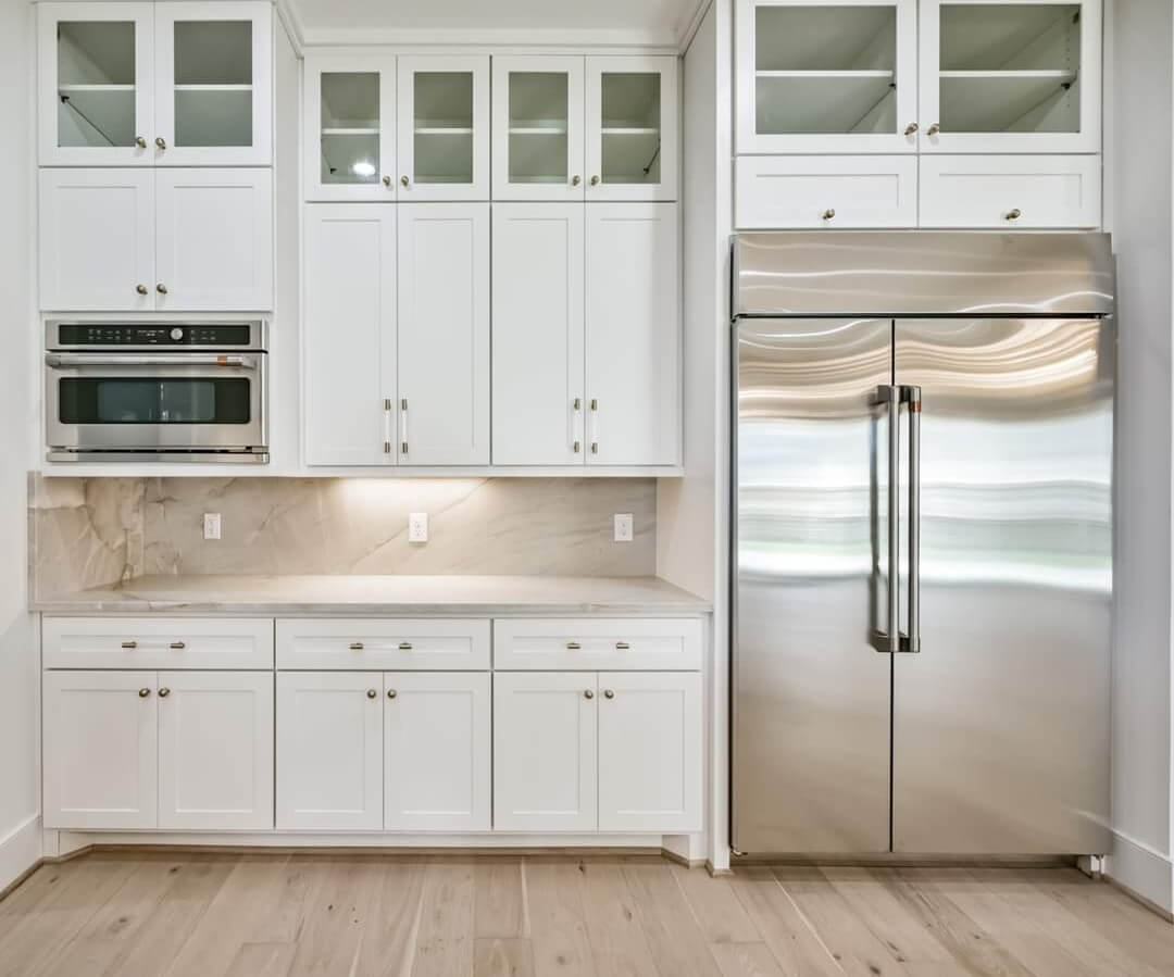 A clean, white pantry area featuring glass-front cabinets, a stainless steel refrigerator, and a marble slab backsplash, completed by Texas Star Painting of Austin.
