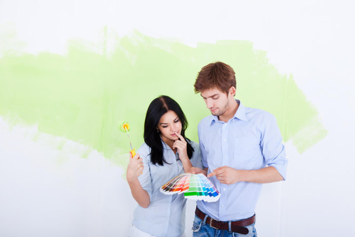 A couple reviewing paint color swatches in front of a partially painted wall, considering the signs it's time to repaint interior walls in their home.