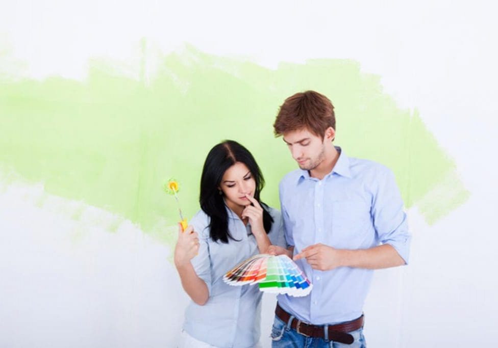 A couple reviewing paint color swatches in front of a partially painted wall, considering the signs it's time to repaint interior walls in their home.